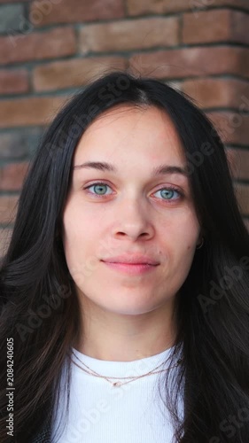 Young woman portrait against brick wall, subtle facial expression shifts and blinking while facing camera, long dark hair, white top, centered vertical framing, direct gaze, still posture.