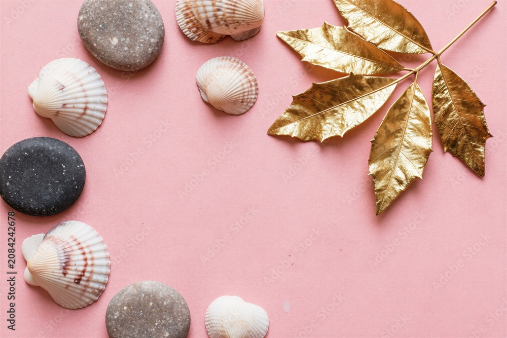 Obraz premium Sea shells and smooth stones arranged on pastel pink surface with golden leaves creating a decorative border. Empty space for text at center.