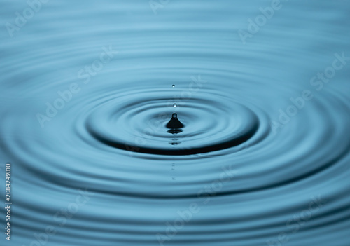 Close up of a blue water drop macro creating concentric ripples on a calm water surface. Abstract liquid background symbolizing purity, freshness, balance and clean water