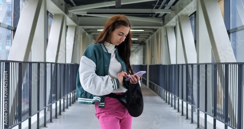 Student texting on glass bridge with varsity jacket and tote, structural beams and long railings frame the scene, checking map and messages while planning route, calm campus atmosphere