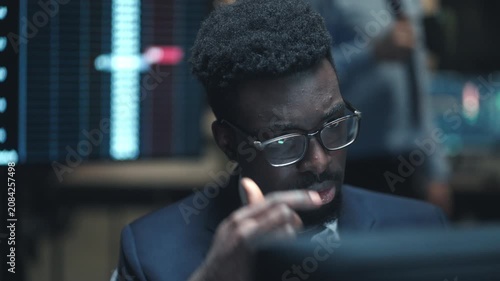 Close up African American male broker wearing glasses and looking at computer screen in dark trading office. Trader analyzing stock market data with financial charts around him late at night.