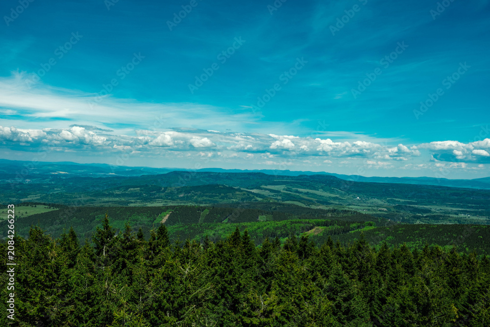 Fototapeta premium Panoramic view from a mountain overlooking a vast landscape of dense evergreen forest, rolling hills, and distant agricultural fields under a blue sky with clouds. Sudetes, Sudety.