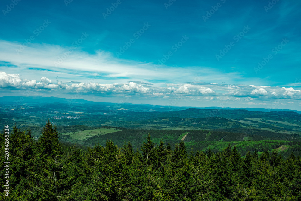Fototapeta premium Panoramic view from a mountain overlooking a vast landscape of dense evergreen forest, rolling hills, and distant agricultural fields under a blue sky with clouds. Sudetes, Sudety.