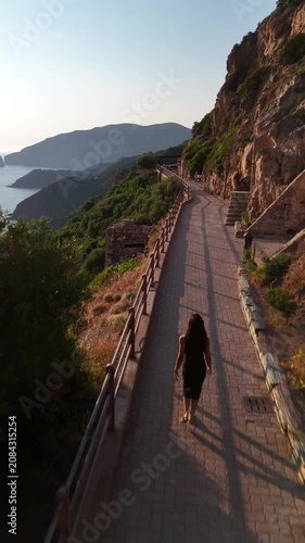 A woman walking along a scenic coastal path near Nebida, filmed from behind as the camera follows her steps against the backdrop of cliffs and Mediterranean sea in south-west Sardinia