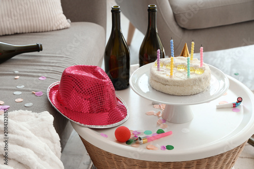 Birthday cake with bottles and party decor on table in messy living room