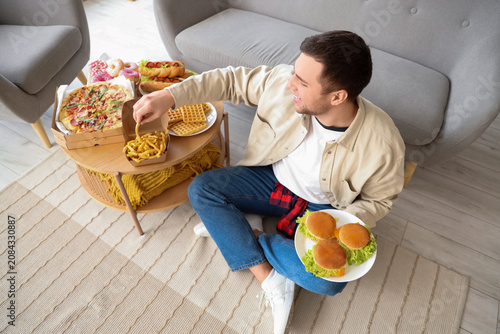 Young man with unhealthy food on table at home. Overeating concept