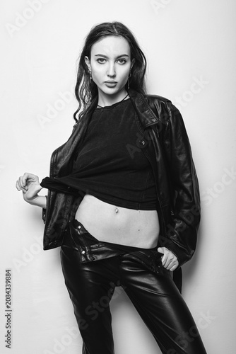 Studio fashion: cool sexy young woman in black leather pants and jacket and t-shirt standing at the wall. Portrait of informal (rock) girl. Black and white
