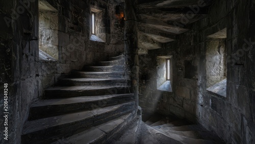 A view of the spiraling helical stone staircase inside a historic medieval castle tower with natural light rays streaming through narrow embrasure windows creating dramatic shadows on the masonry