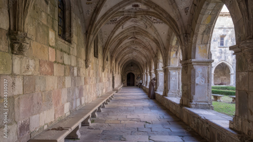 Obraz premium A robed monk bows his head in prayer inside the vaulted stone corridor of a historic monastic cloister featuring weathered masonry arches that overlook a serene green courtyard at dawn light