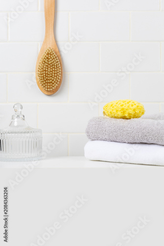 Minimalist bathroom product mockup background with copy space in foreground. Hanging wooden body brush, yellow sea sponge, glass jar, and stacked towels against white subway tile wall.
