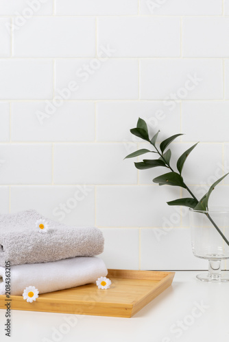 Minimalist spa product mockup background with white subway tile wall. Stacked soft towels on a wooden tray decorated with small daisy flowers and a green leaf branch in glass.