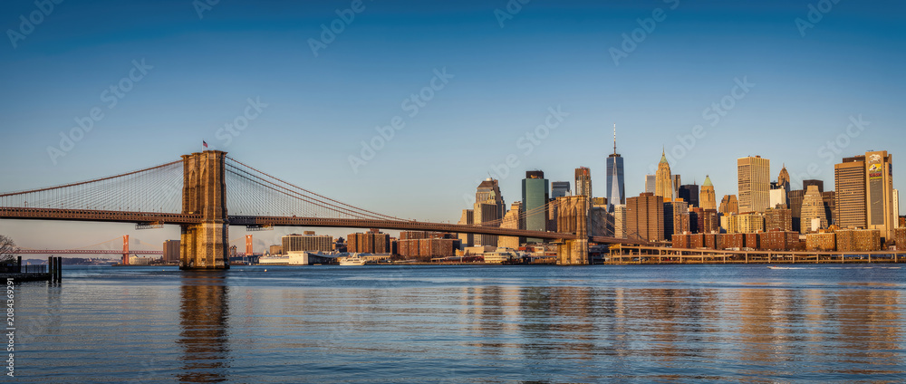 Fototapeta premium Iconic Brooklyn Bridge and Lower Manhattan Skyline at Golden Hour, New York City. Panoramic view of historic architecture, skyscrapers, and East River reflections under a clear blue sky.