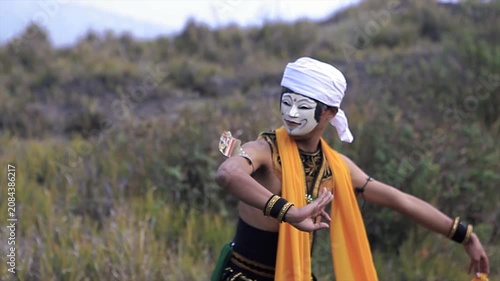 Traditional Indonesian dancer in a white mask and yellow sash performing on a grassy hill, 4K video.