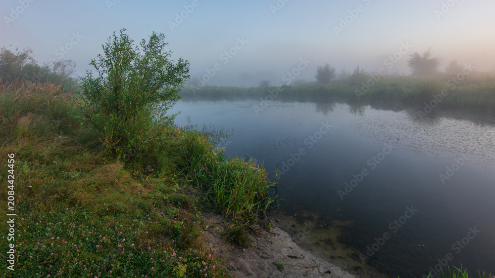 Obraz premium A serene, misty morning landscape features a calm river reflecting the soft sky, surrounded by lush green banks and hazy trees