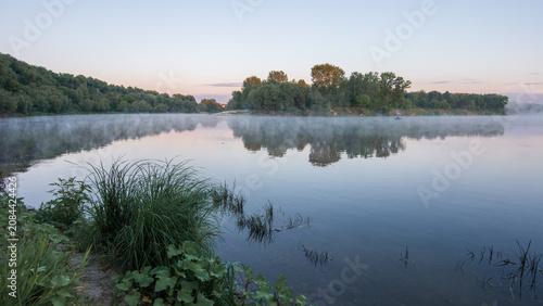Fotografie A serene river flows calmly at dawn, reflecting lush green trees and misty banks