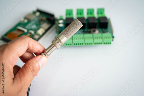 Fotografía Close-up of an industrial temperature and humidity sensor held by hand with electronic controller board in the background, representing IoT, environmental monitoring, smart automation systems