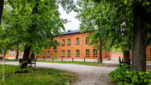 Historic red brick building with green windows, located in a green park with trees and a gravel path in the foreground, characteristic architecture of the Ksiezny Mlyn district in Lodz, daylight.