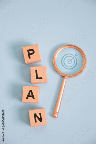 Business planning concept with wooden letter blocks spelling PLAN and magnifying glass highlighting target strategy on blue background, symbolizing goal setting, project management, vision, success.