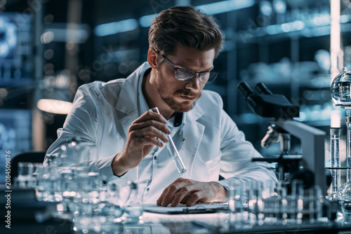 Scientist in lab coat examines test tube with focused expression and glasses