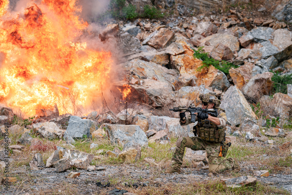 Fototapeta premium Soldier takes cover behind rocks during training near explosion in rocky area