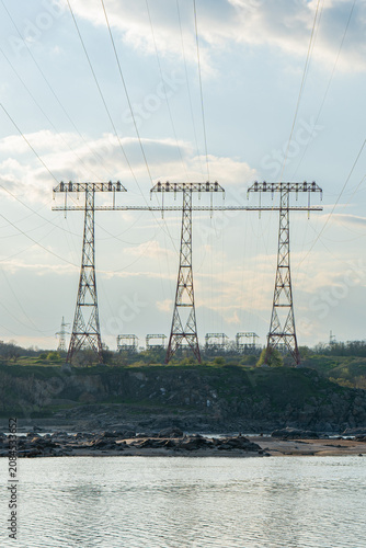 Tall electricity pylons stand in a row across a rocky landscape near water