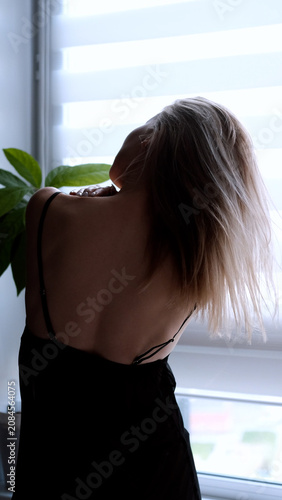 A sexy beautiful woman in a black erotic negligee stands near the window in the bedroom.