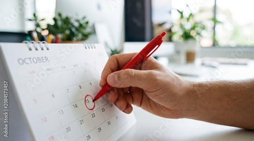 Close-Up View of Bare Hand Holding Vibrant Red Pen Circling Date on White Desk Calendar, Highlighting Time Management Organization, Workplace Productivity, and Modern Office Business Planning Concepts