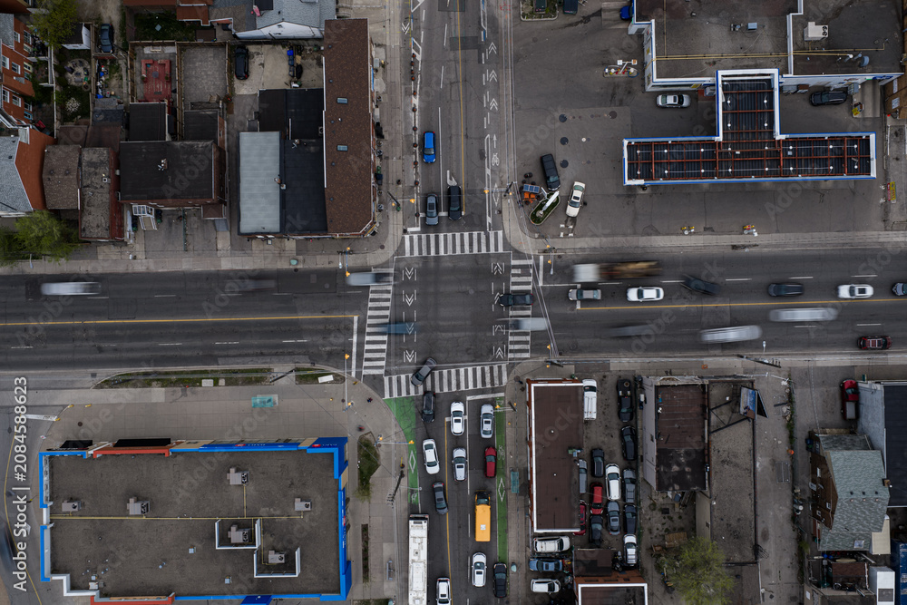 Obraz premium Aerial top down view of busy Toronto city intersection with moving cars, marked crosswalks, bike lanes, rooftops, parking areas, and dense urban blocks under overcast light