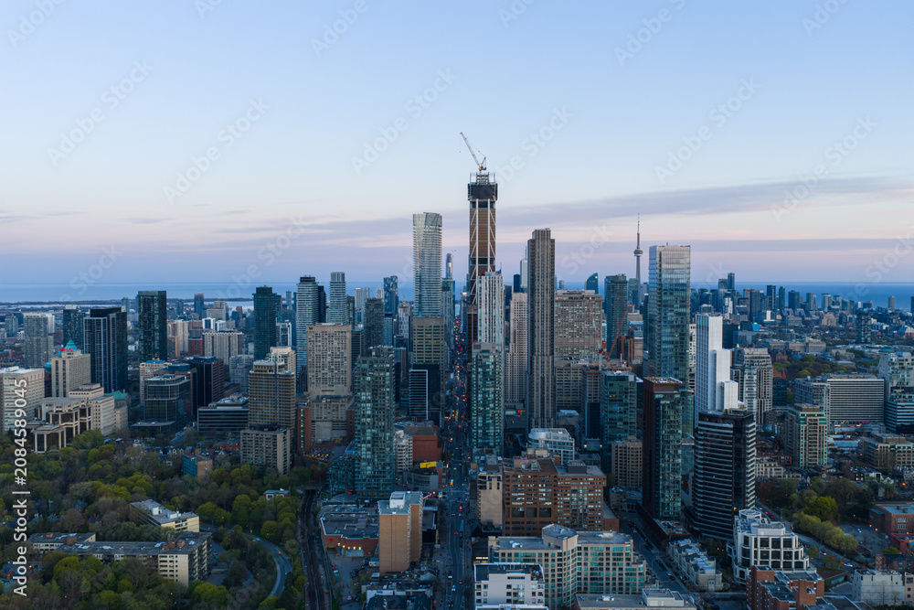Obraz premium Toronto skyline at dusk with high rise towers, construction crane, distant lake horizon, layered downtown buildings, soft evening light, clear elevated city view over urban core