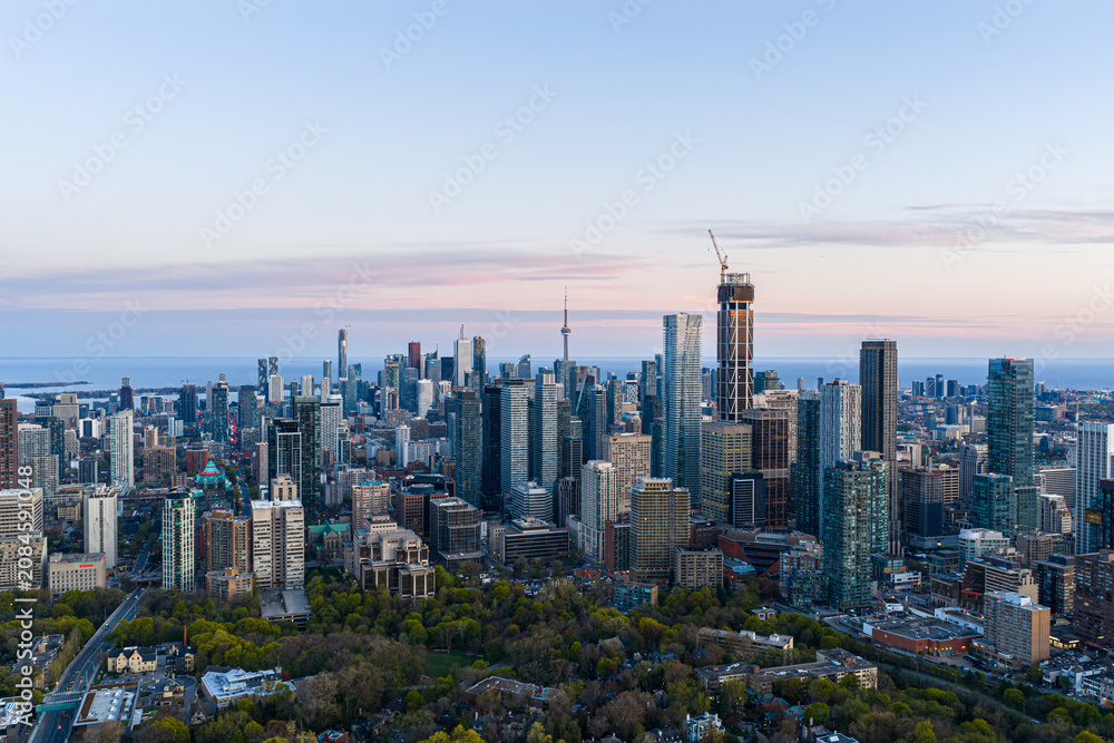 Obraz premium Toronto skyline at dusk with dense downtown towers, CN Tower, rising high-rises, construction crane, lake horizon, pastel evening sky, urban neighborhoods, and tree-lined foreground
