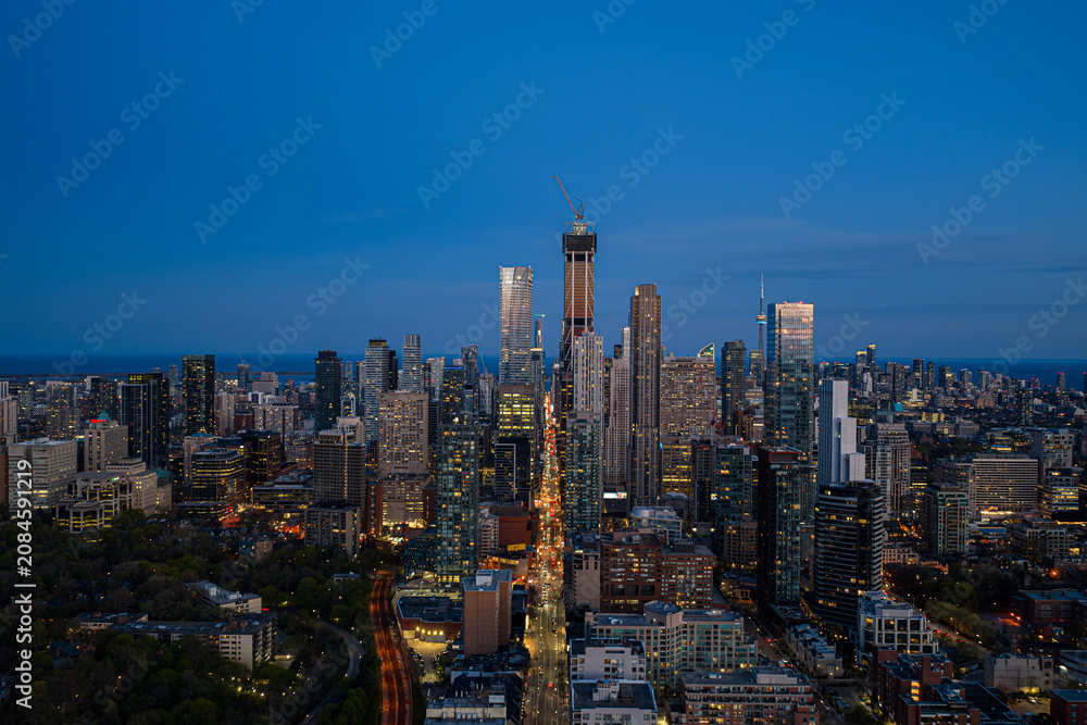 Obraz premium Dusk aerial view over Toronto skyline with illuminated high-rise towers, dense downtown streets, and bright blue evening sky stretching above Lake Ontario
