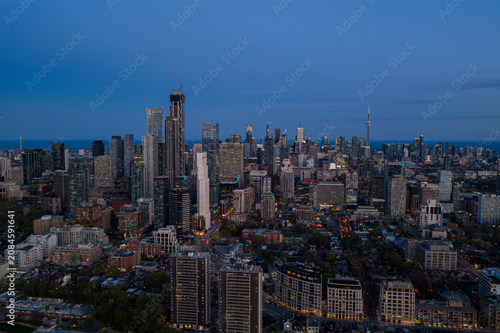 Obraz premium Toronto skyline at dusk with illuminated high-rise towers, construction crane, and wide urban panorama under deep blue evening sky above dense downtown district near lake horizon