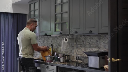 A wide commercial shot of a happy adult man moving rhythmically and dancing while washing dishes in a modern kitchen.