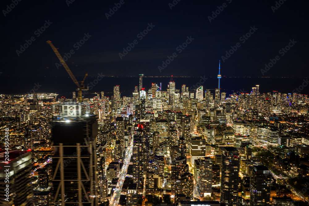 Obraz premium Night aerial view over downtown Toronto with illuminated high rise towers, construction crane on high rise building foreground, dense street grid, and tall landmark tower glowing against dark sky