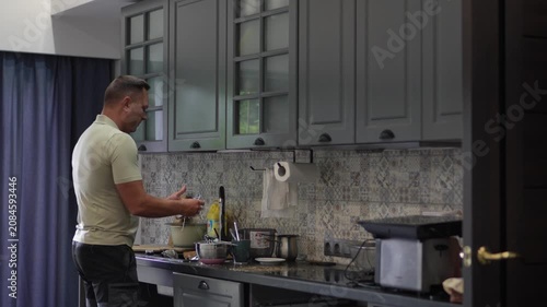 A happy middle-aged man enthusiastically singing into a metal spoon like a microphone while doing chores in a modern kitchen.