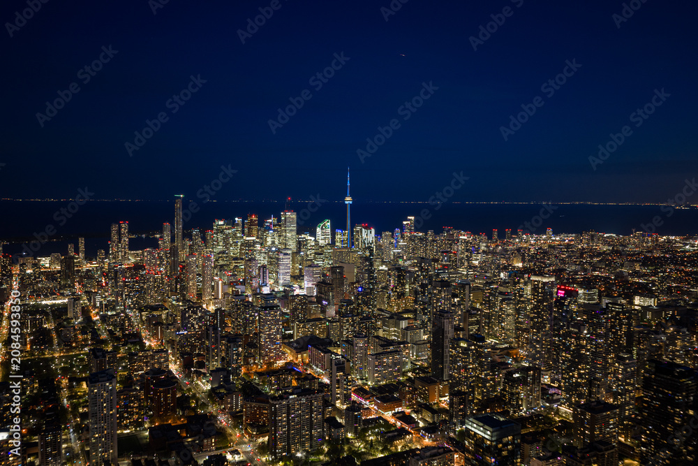 Obraz premium Toronto night skyline glowing above dense downtown towers with city lights, illuminated streets, distant lake horizon, and iconic central tower seen from high aerial viewpoint