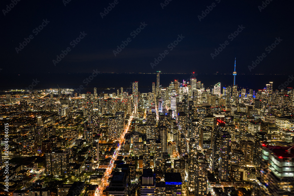 Obraz premium Toronto skyline glowing at night with dense downtown towers, illuminated streets, dark lake horizon, and iconic city lights viewed from high aerial perspective over Canada
