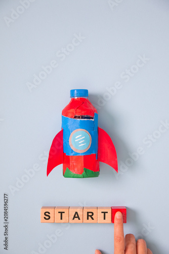 Creative startup concept with handmade rocket, wooden START blocks and finger pressing launch button on blue background, symbolizing business growth, innovation, entrepreneurship, motivation, success.