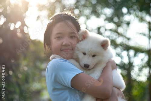 A happy Asian girl gently hugging a cute fluffy white puppy, smiling, outdoor natural light, friendship and love concept.