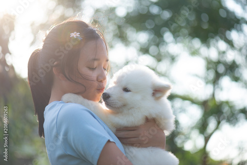 A happy Asian girl gently hugging and kissing a cute fluffy white puppy, outdoor natural light, friendship and love concept.