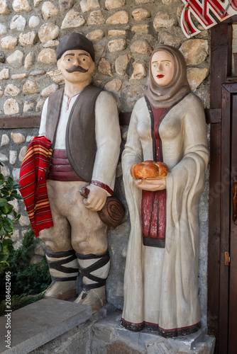 Two life-sized painted statues in traditional Bulgarian folk attire stand outside a stone building, representing local culture and customs with rustic charm in Bansko, Bulgaria.