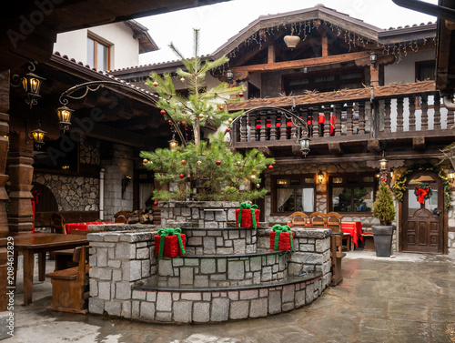 A cozy stone and wood courtyard of a traditional Bulgarian inn, decorated for the holidays with a Christmas tree and festive gift boxes in Bansko, Bulgaria.