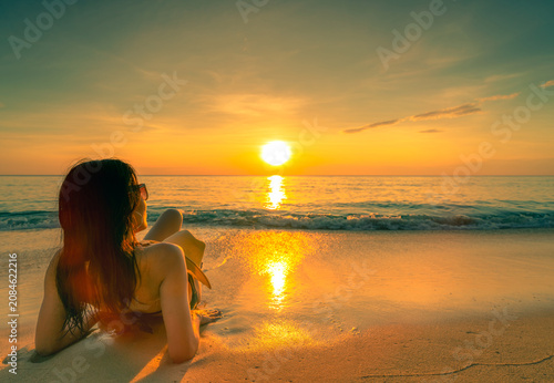 Fotografie Woman relaxing on a tropical sandy beach while watching sunset over calm ocean