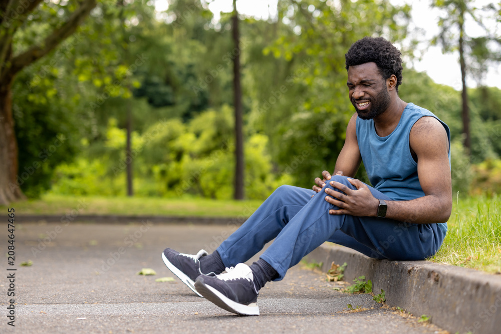 Obraz premium Runner sitting on a curb gripping his knee, feeling intense pain after a sport accident, highlighting themes of health issues, urgency, and injury prevention in athletics