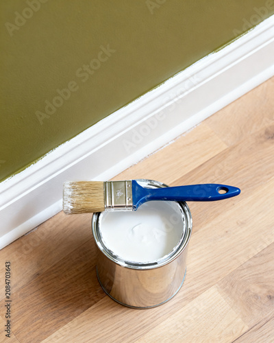 A paint brush resting on a metal tin of white wall paint placed on a wooden floor near a freshly painted green interior wall baseboard