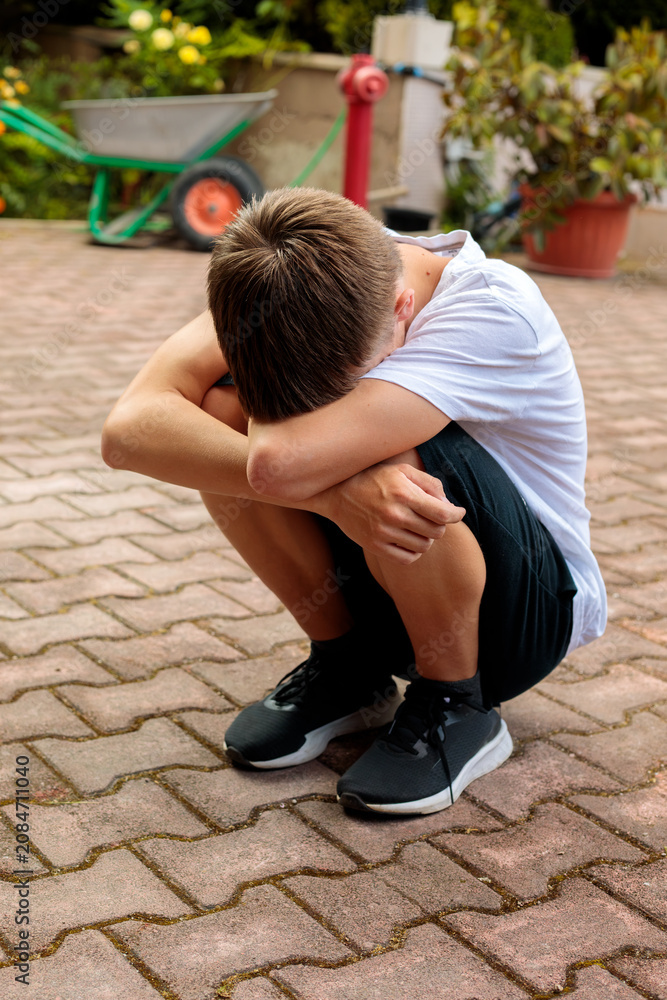 Obraz premium Sad teenage boy sitting on pavement with head on folded arms, expressing loneliness, stress, emotional pain and depression in an outdoor urban setting alone