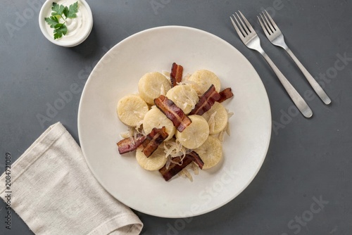 Plate of Strapacky garnished with bacon, served with creamy white sauce and parsley on plain grey background. Concept of potato dumplings with sauerkraut bacon.