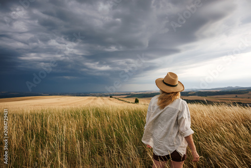 Worried female farmer looking at approaching storm clouds over agricultural field. Summer weather and impact climate change on agriculture concept