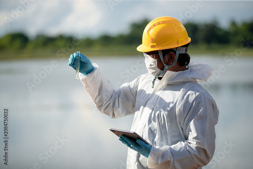 Ecologist examining water quality with flask and tablet near natural water source in protective gear