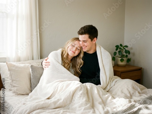 Young couple sitting together in bed, wrapped in a cozy white blanket.